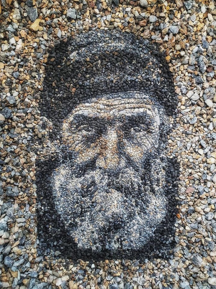 A pebble portrait titled "Fisherman" by Justin Bateman, featuring the face of an older man with a detailed beard and a rugged hat. Created using carefully selected stones in shades of black, gray, and white, the portrait showcases Bateman's talent for capturing intricate facial expressions and textures. The piece is set on a gravel background, blending seamlessly into its natural surroundings in Chiang Mai, Thailand.