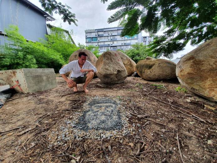 Justin Bateman crouches next to his pebble portrait titled "Fisherman" in a natural outdoor setting in Chiang Mai, Thailand. The artwork features a bearded man’s face, intricately crafted from stones of varying colors and textures. Surrounded by large boulders and lush greenery, the setting contrasts with an urban building in the background, emphasizing the temporary and harmonious nature of Bateman’s art.