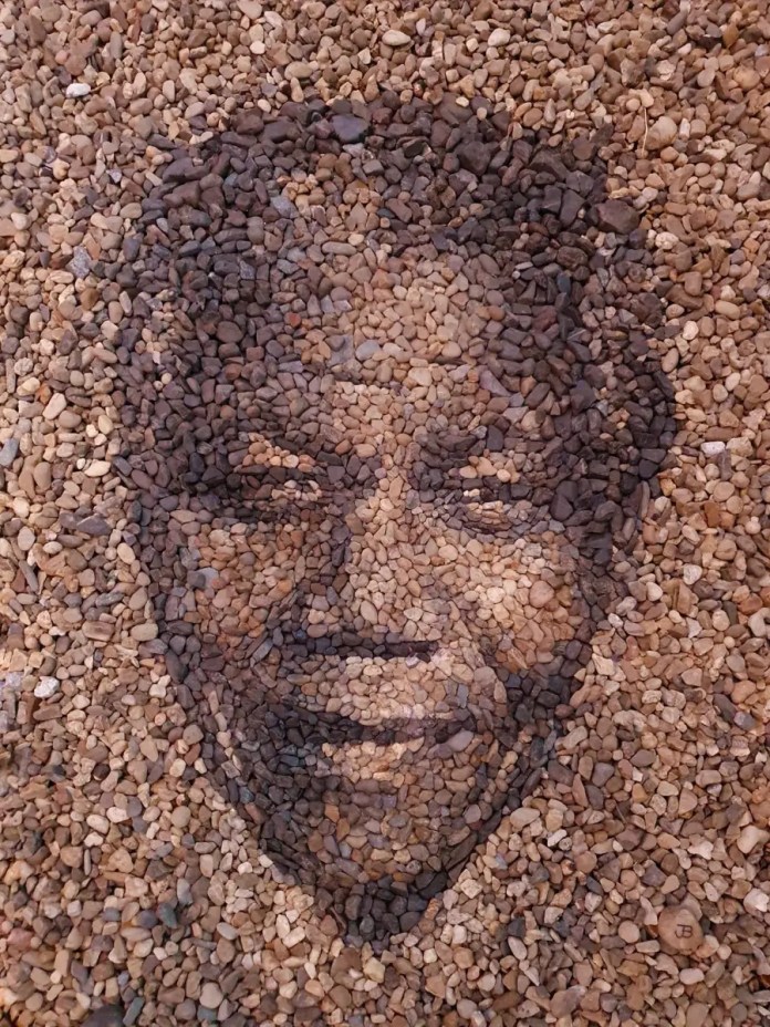 A stone and pebble portrait titled "Mandela Mandala" by Justin Bateman, featuring a warm depiction of Nelson Mandela. The artwork is crafted using earthy brown and tan stones, arranged to highlight the contours of Mandela’s face and his gentle expression. The pebbles’ natural tones enhance the portrait's depth, symbolizing Mandela’s enduring legacy and connection to the earth. Created in Chiang Mai, Thailand, this piece reflects Bateman's skill in combining simplicity and profound storytelling.