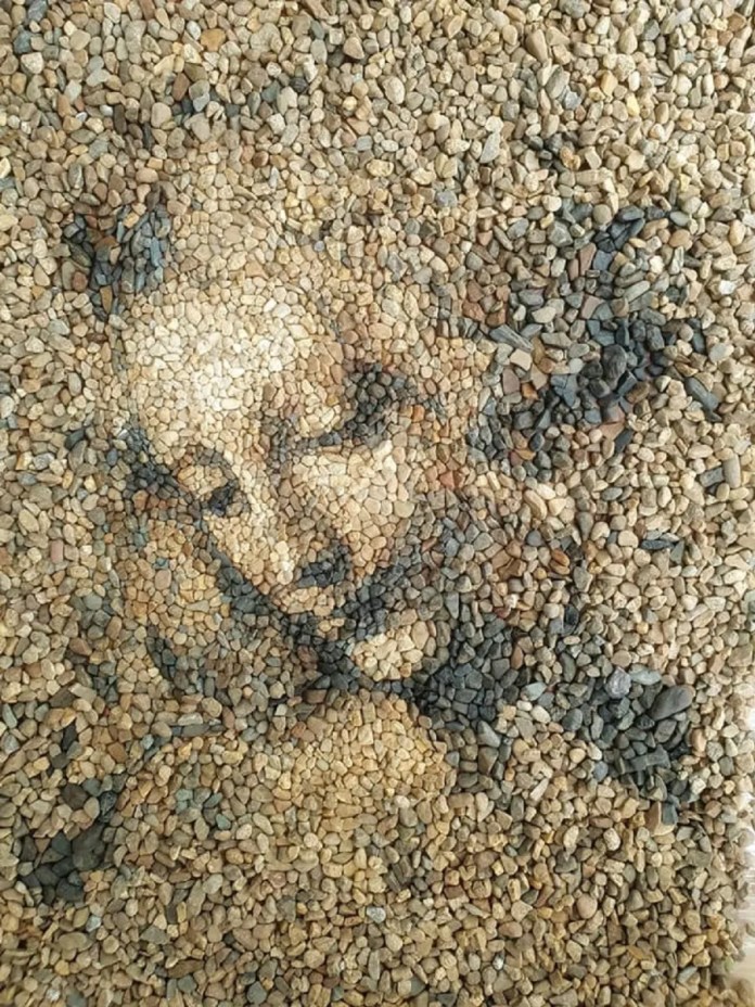 A delicate stone and pebble portrait titled "La Scapigliata" by Justin Bateman, inspired by Leonardo da Vinci’s unfinished masterpiece. The artwork uses a blend of tan, beige, and dark pebbles to recreate the serene expression and flowing features of the subject. The intricate design captures both the softness and depth of the original artwork, blending seamlessly into the natural gravel setting. Created in Chiang Mai, Thailand, this piece highlights Bateman’s ability to reinterpret classical art using ephemeral materials.