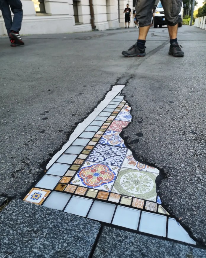 Street crack in Belgrade filled with a mosaic of square tiles and decorative ceramics in blue, orange, and green; feet of two people nearby emphasize the scale.