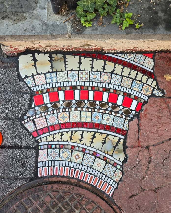 Curved mosaic repair in Ankara featuring red, white, and beige tiles arranged in layered arcs beside a manhole and ivy-covered wall.
