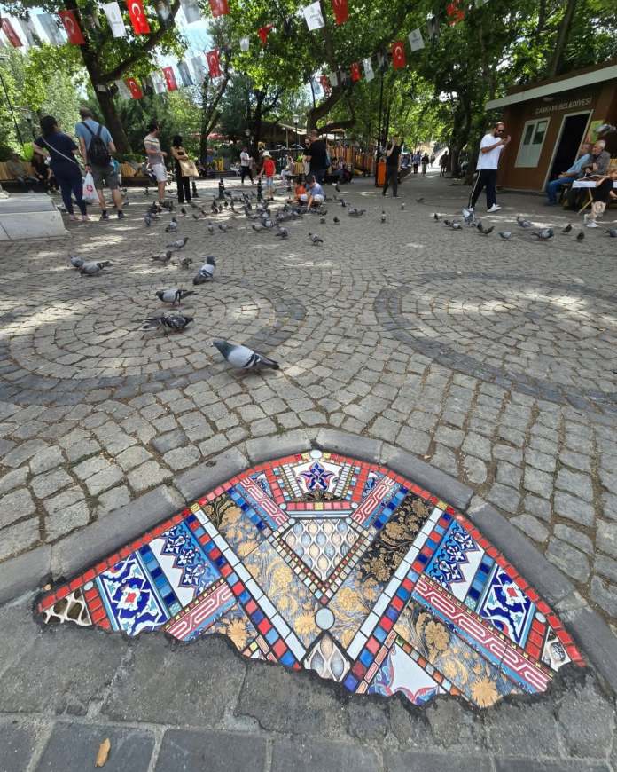 Large mosaic in Ankara plaza shaped like a half-moon, embedded in cobblestones, with intricate red, blue, and gold tilework surrounded by pigeons and people.