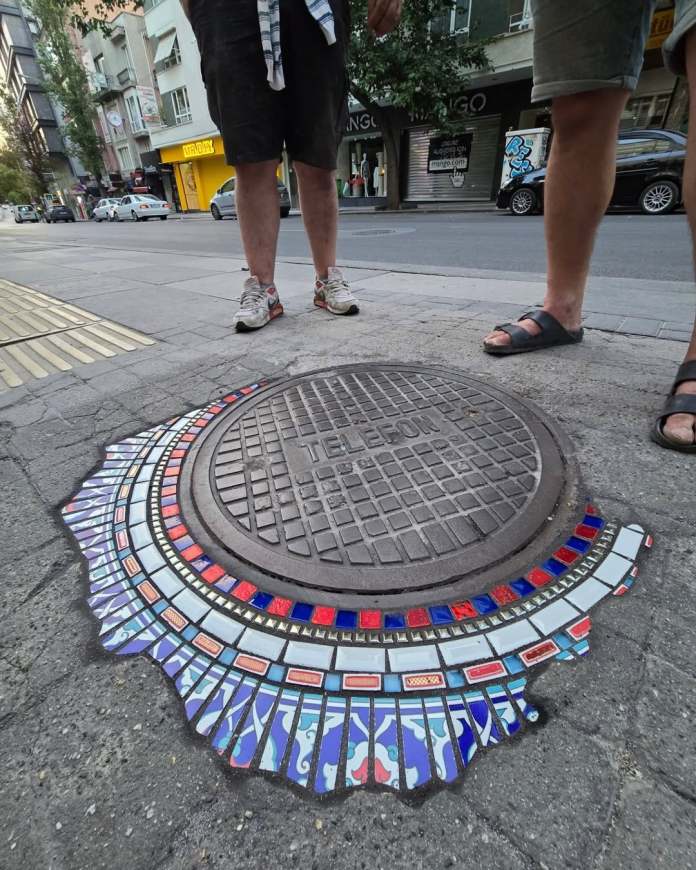 Ankara street manhole cover encircled with a decorative mosaic of white, red, and blue tiles with a scalloped edge.