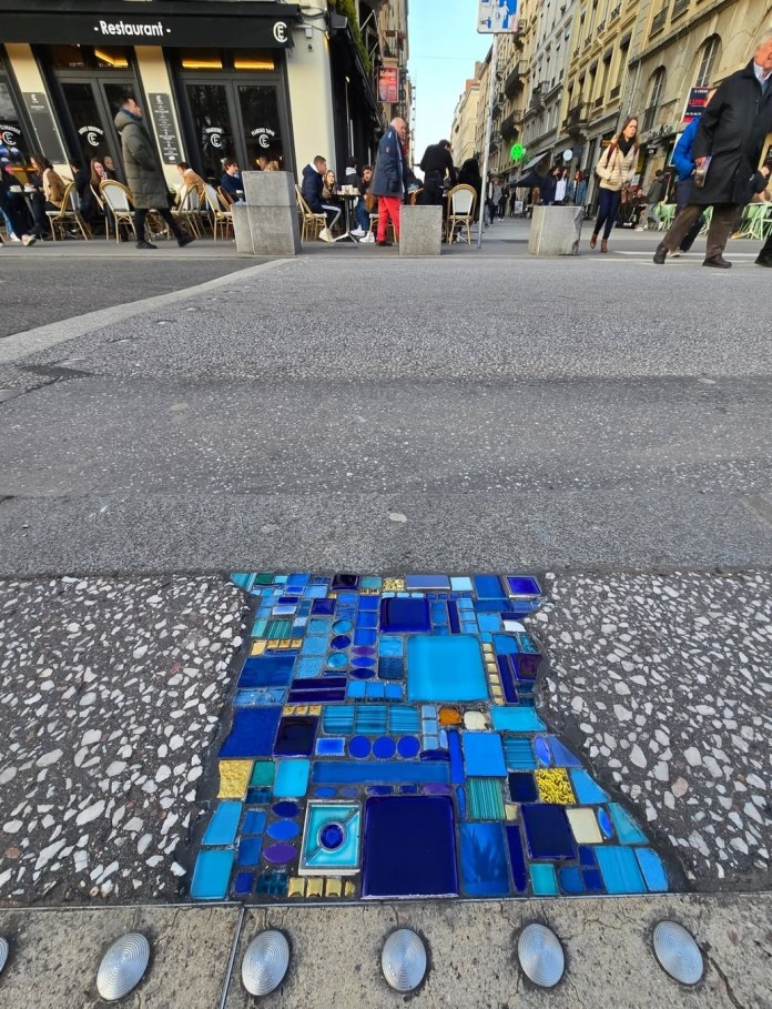 Urban street crack in Lyon filled with blue glass mosaic tiles in a grid pattern next to terrazzo pavement and concrete.