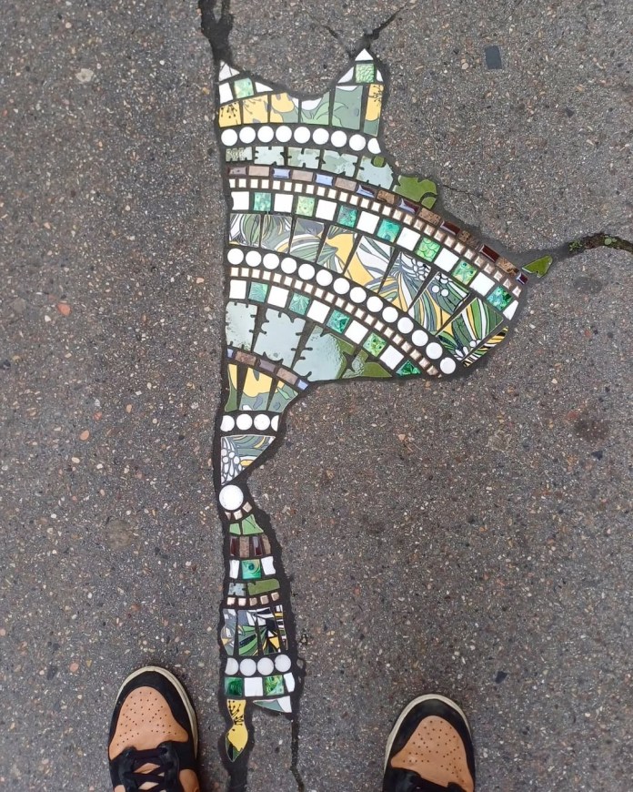 Mosaic filling a jagged crack in the street in Lyon, with layered curved lines of green, yellow, and white tiles, surrounded by speckled asphalt and brown sneakers.