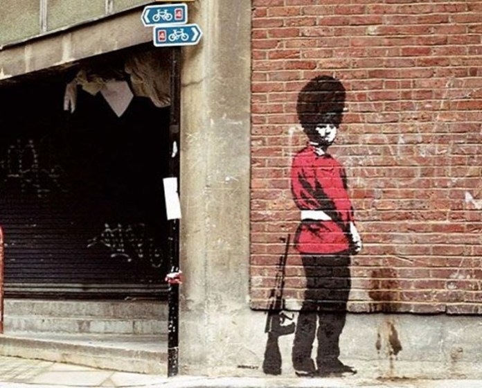 Stencil artwork of a Queen’s Guard in red and black uniform facing a wall, suggesting he is urinating. The streak on the brick wall adds realism. Painted directly onto a London street corner.