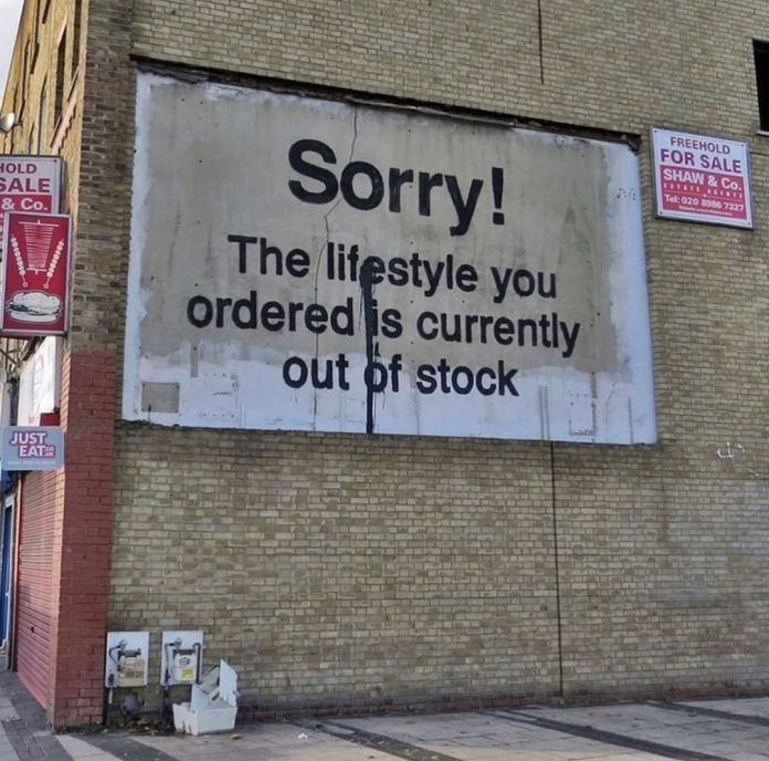 Banksy artwork on a whitewashed billboard reading “Sorry! The lifestyle you ordered is currently out of stock.” Positioned high on a brick building wall.
