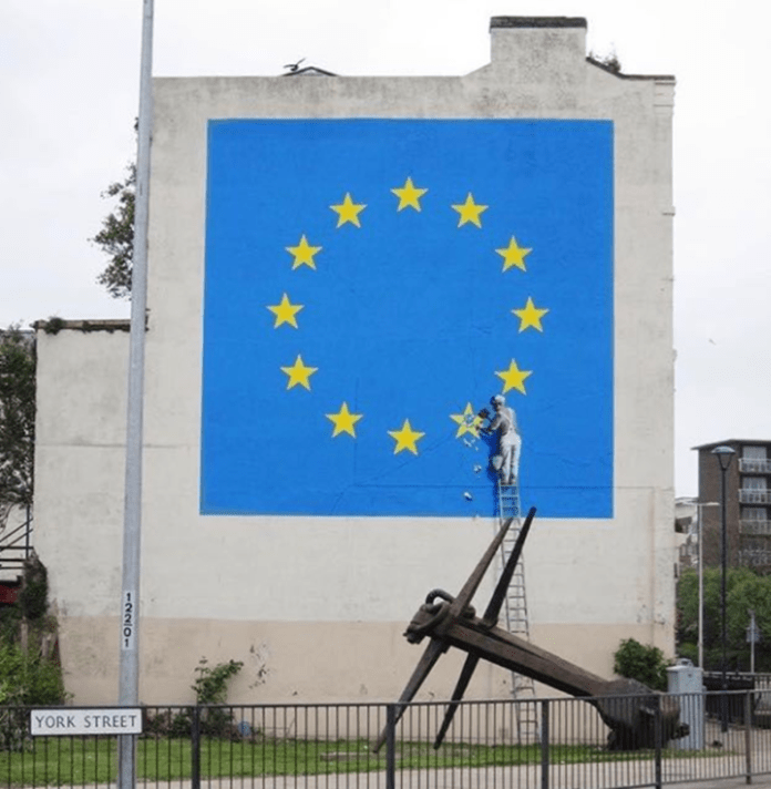 A worker on a ladder chips away one star from the EU flag. Created during Brexit negotiations, the mural questions whether Britain’s exit is liberation—or self-destruction.