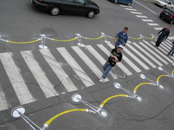 Pedestrians crossing a street where the crosswalk is painted to resemble a red carpet with stanchions, a creative piece of street art by Roadsworth.