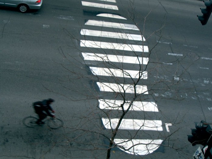 A large footprint design painted on a crosswalk by Roadsworth, blending with the street lines in an urban setting.