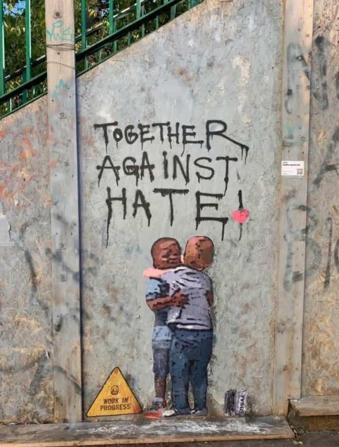 A street art mural by TVBOY featuring two children embracing against a rough, gray wall. Above them, the message “TOGETHER AGAINST HATE!” is painted in bold, dripping black letters, with a small red heart accentuating the word “HATE.” A “WORK IN PROGRESS” sign sits at the bottom, emphasizing unity as an ongoing effort. The mural is painted on a textured surface, surrounded by graffiti, reinforcing its raw and socially conscious message.