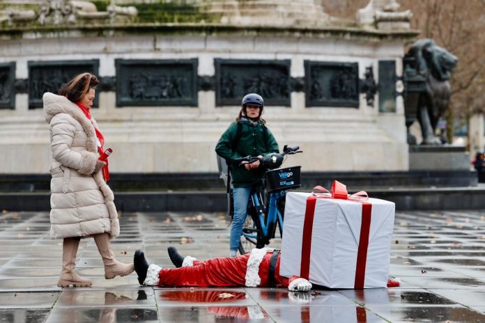 A close-up of James Colomina's Santa Claus installation in Paris. The sculpture shows Santa Claus lying flat on a wet, reflective pavement with a large white gift box, tied with a red ribbon, replacing his head. In the background, pedestrians walk by under cloudy skies, adding a sense of motion and contrast to the stillness of the artwork.