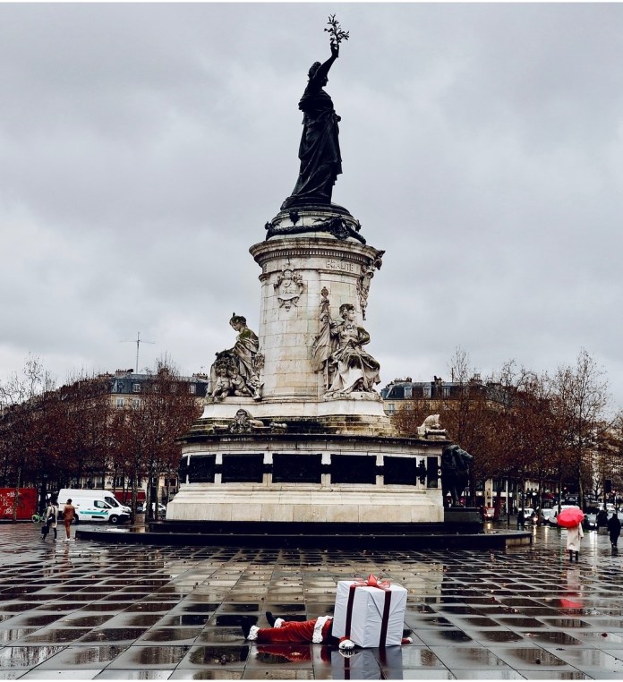 James Colomina's Santa Claus sculpture in front of the Place de la République monument in Paris. The scene shows Santa lying flat on the reflective pavement with a large gift box as his head, set against the towering statue symbolizing liberty. The overcast sky and sparse pedestrians add to the contemplative atmosphere.
