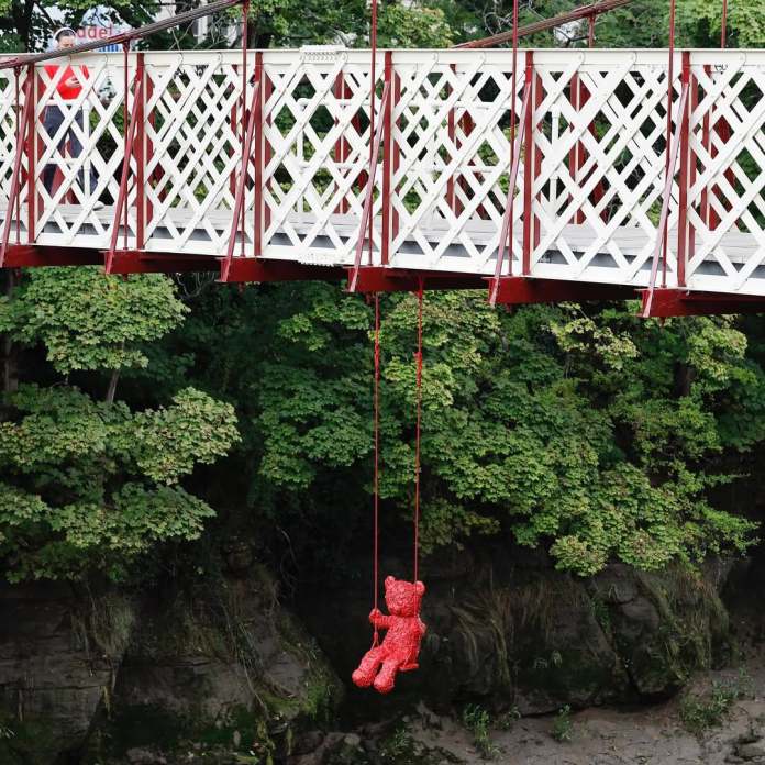 A red sculpture of a teddy bear by James Colomina swings beneath the Gaol Ferry Bridge in an urban setting surrounded by lush greenery. The bear hangs on red ropes from the intricately designed white and red bridge, creating a striking contrast with the natural backdrop. A pedestrian can be seen on the bridge, adding a sense of scale and human presence to the scene.