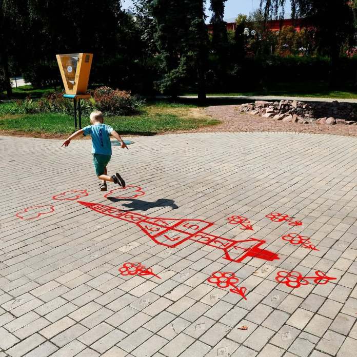 A child hops through La Marelle, a red-painted hopscotch artwork by James Colomina in a public park. The design creatively transforms the traditional hopscotch game into the shape of a dove surrounded by flowers, blending playfulness with a message of peace. The scene is set against a background of trees and park features, capturing a moment of joy and reflection.