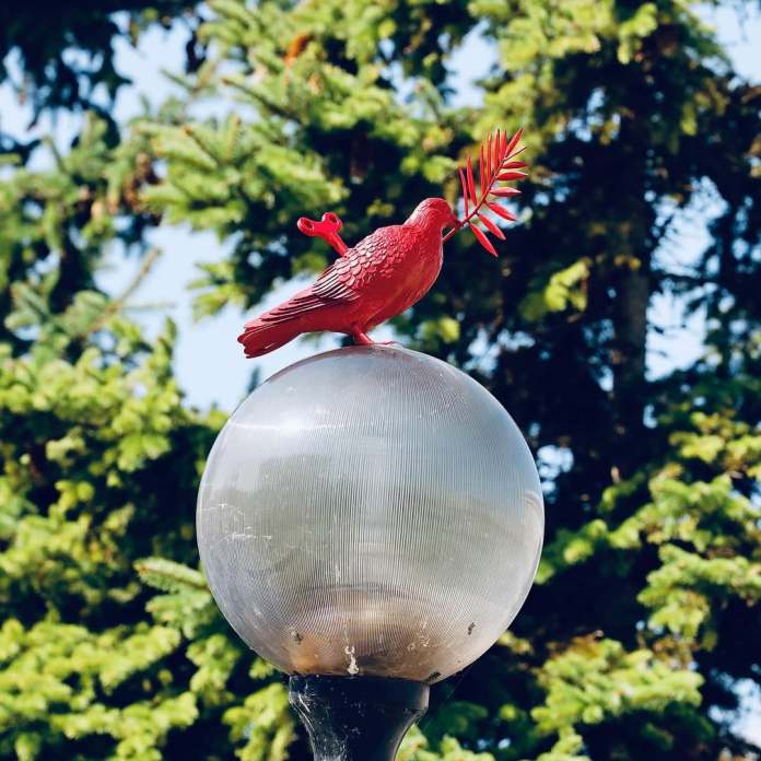 A red sculpture of a dove by James Colomina perches atop a round streetlamp, holding an olive branch in its beak. The bright red of the dove contrasts against the green trees in the background, symbolizing peace and hope through this striking public artwork titled La Colombe.