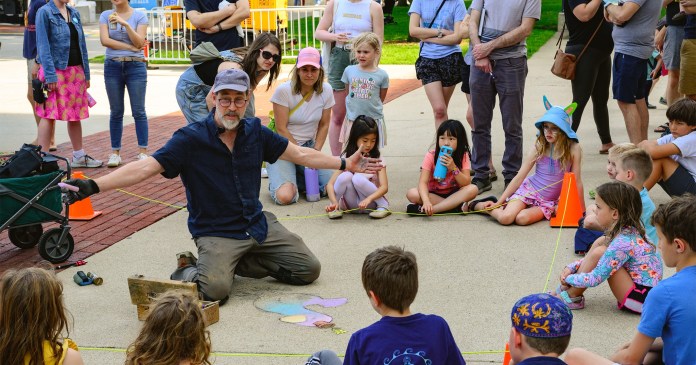 David Zinn conducting a hands-on chalk art class outdoors, surrounded by an attentive group of children and adults. Zinn is kneeling on the pavement with open arms, explaining his process as colorful chalk materials and an unfinished whimsical drawing of a bunny are displayed in front of him. The children sit in a semi-circle, fully engaged, while adults stand in the background observing the creative session. The scene radiates community and creativity, capturing the joy of interactive art-making.