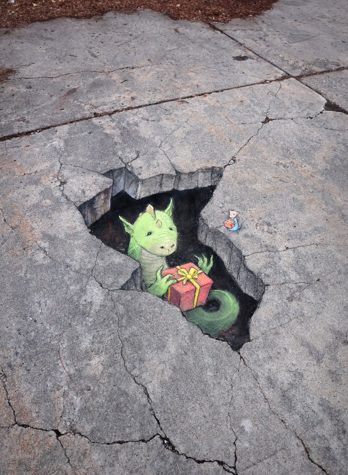 A vibrant chalk art creation by David Zinn, featuring a friendly green dragon emerging from a crack in the pavement. The dragon holds a red gift box with a yellow ribbon, its expression warm and inviting. To the right, a tiny chalk-drawn mouse observes the scene while holding a matching gift. The artwork creatively integrates with the cracked surface, making the scene appear three-dimensional and magical.