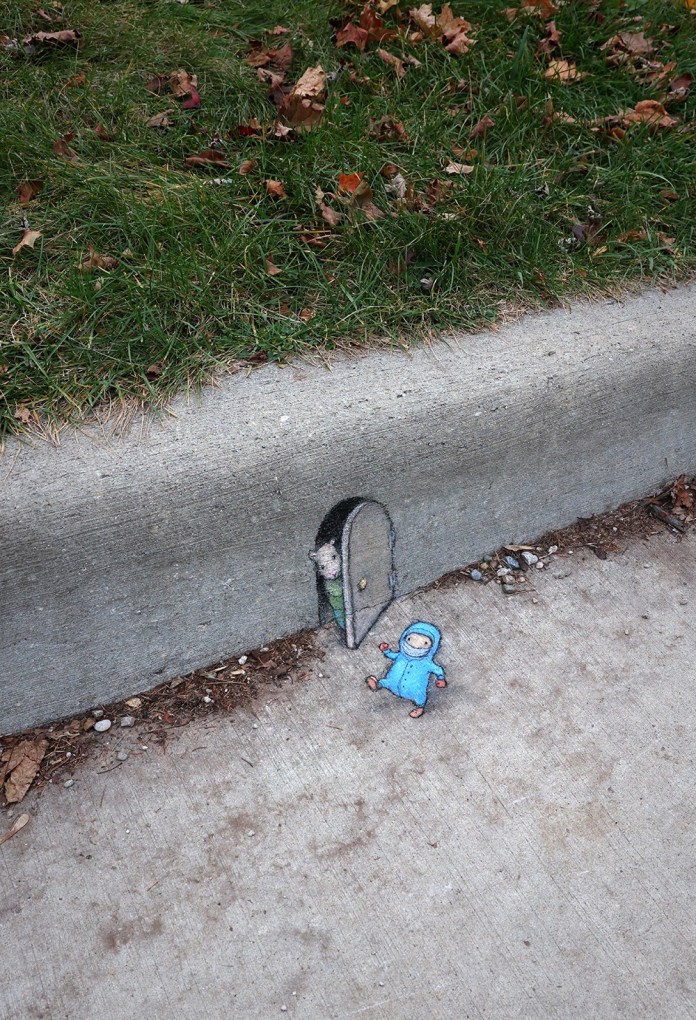 A playful chalk art piece by David Zinn depicting a small arched doorway drawn into a concrete curb. A tiny mouse peeks out from behind the partially opened door, while a small character in a blue snowsuit lies on the sidewalk outside, appearing unfazed by the cold. The scene cleverly blends with the curb and adjacent grass, creating a whimsical winter-themed vignette.