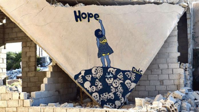 A poignant mural painted on the triangular wall of a partially destroyed building in Darayya, Syria, created by the artist Abu Malik al-Shami. The artwork depicts a young girl in a yellow-polka-dot blue dress reaching up to write the word "Hope" in yellow letters. She stands atop a pile of skulls, symbolizing the devastating toll of war, while the crumbling building around the mural reflects the harsh realities of conflict. The artist’s signature, "DMC," is visible in the corner, alongside the Arabic name for Darayya. This mural is a powerful statement on resilience and optimism amidst destruction.