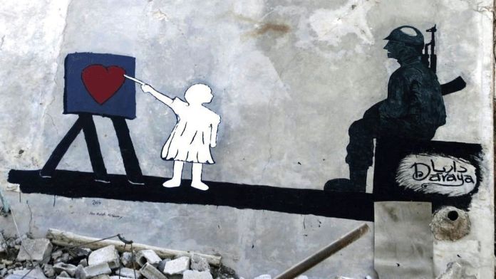 A thought-provoking mural by Abu Malik al-Shami on the wall of a war-damaged building in Darayya, Syria. The artwork features the silhouette of a soldier with a rifle slung over his back, sitting contemplatively on one side. Opposite him, a child in a white dress stands, painting a red heart on a canvas. The stark contrast between the soldier and the child's act of creation highlights themes of war and peace. The signature "Daraya" is inscribed on the wall, emphasizing the location’s context within the Syrian conflict. Rubble surrounds the scene, reinforcing the mural's message of hope and humanity amidst destruction.