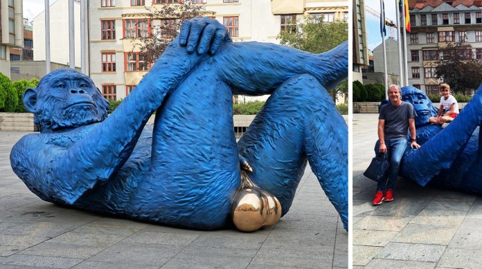 A striking sculpture titled 'King Kong Balls' by Denis Defrancesco, featuring a blue gorilla with exaggerated golden testicles lying casually in an urban plaza. The left side highlights the full sculpture, while the right shows public interaction, with an adult and child sitting on its arm and leg. The vibrant blue and gold colors contrast against a backdrop of modern buildings, trees, and flagpoles, capturing both humor and engagement in public art.