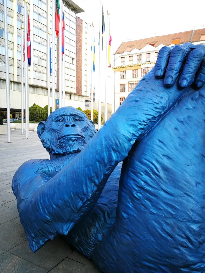 Close-up view of the "King Kong Balls" sculpture by Denis Defrancesco, highlighting the blue gorilla's face and hands as it lies on its back with legs crossed. The detailed texture of the bronze sculpture emphasizes its artistic craftsmanship. In the background, a row of flagpoles with international flags, modern buildings with glass facades, and greenery add context to the sculpture's placement in a public plaza.