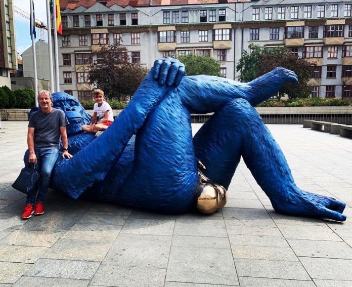 The "King Kong Balls" sculpture by Denis Defrancesco, a bold bronze artwork of a blue gorilla lying on its back with legs crossed and prominent golden testicles. Two people, an adult and a child, interact with the sculpture, sitting on its arm and leg. The scene is set in an urban plaza with modern buildings, trees, and flagpoles in the background, highlighting the playful engagement between the art and its audience.