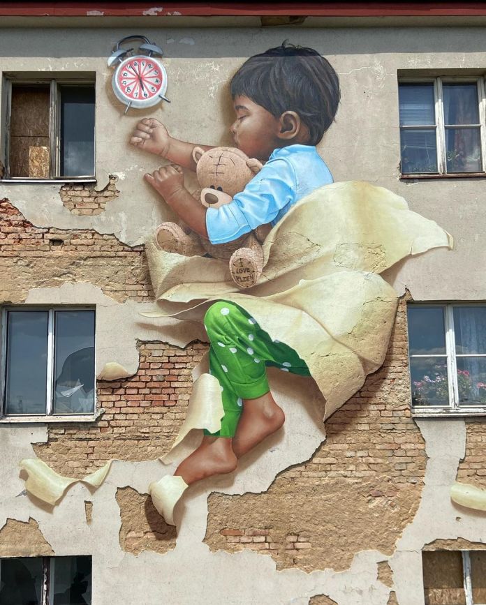 mural by Chemis in Plzeň, Czech Republic, depicting a young child peacefully asleep while clutching a teddy bear. The artwork masterfully integrates the peeling façade of the building into the scene, making it appear as if the child is tucked into a blanket of torn plaster. An alarm clock floats above, adding a dreamlike quality. The teddy bear has "I Love Plzen" inscribed on its foot, subtly tying the mural to its location.