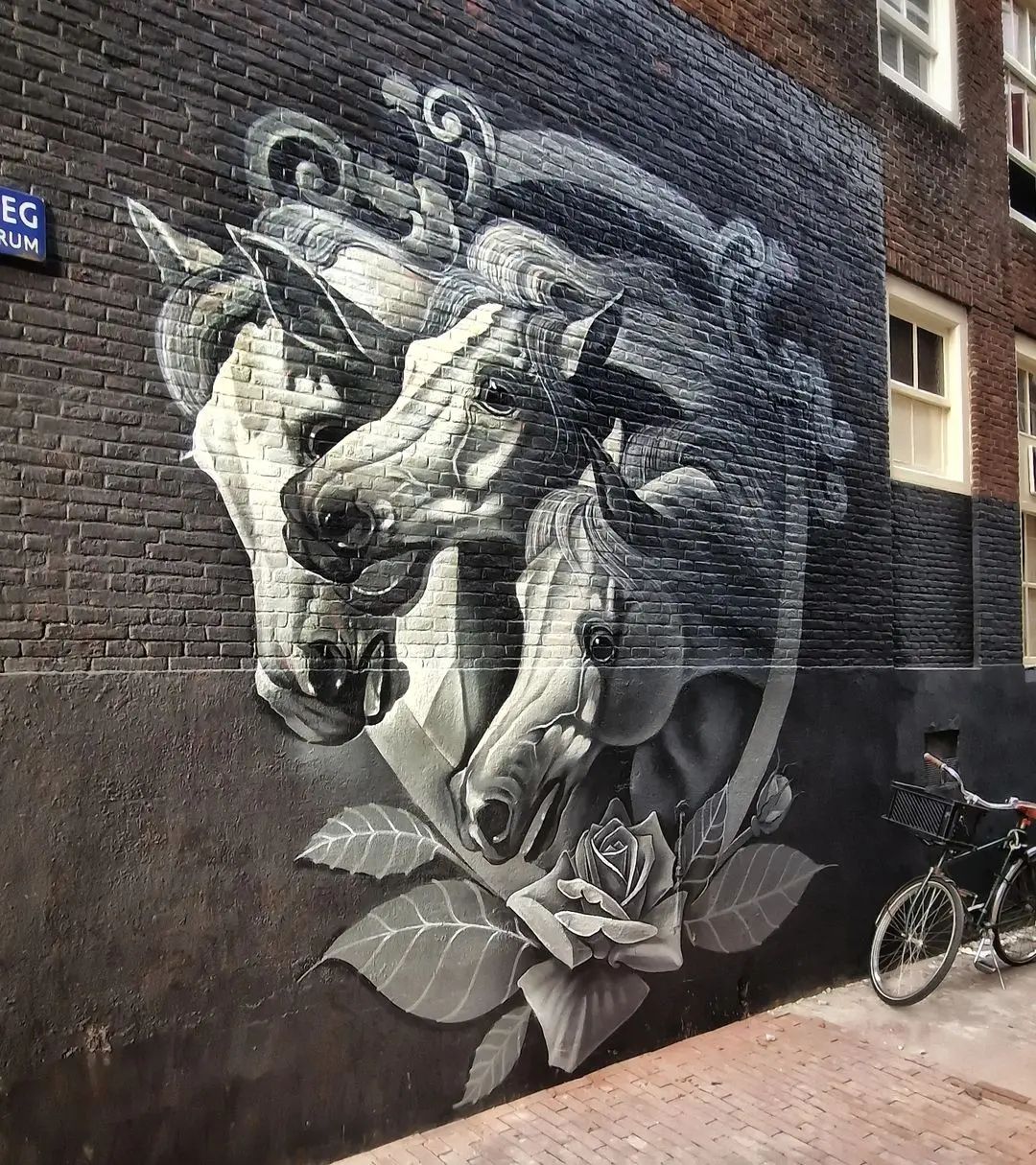 Black-and-white mural by Łukasz Kieł in Amsterdam, Netherlands, showing three realistic horse heads surrounded by roses and leaves, painted on a brick wall with fine gradient shading.