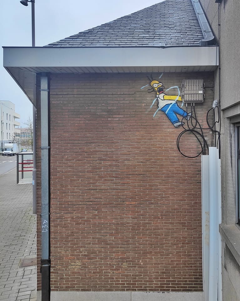 A humorous street art piece by Oakoak featuring Homer Simpson from The Simpsons being electrocuted. The artwork is cleverly positioned on a brick wall next to exposed electrical wires and a utility box, making it appear as though Homer is being shocked by the wiring. The cartoonish depiction includes electric sparks drawn around Homer for a dramatic and comedic effect. This playful integration of the environment into the art is a hallmark of Oakoak's creative style.