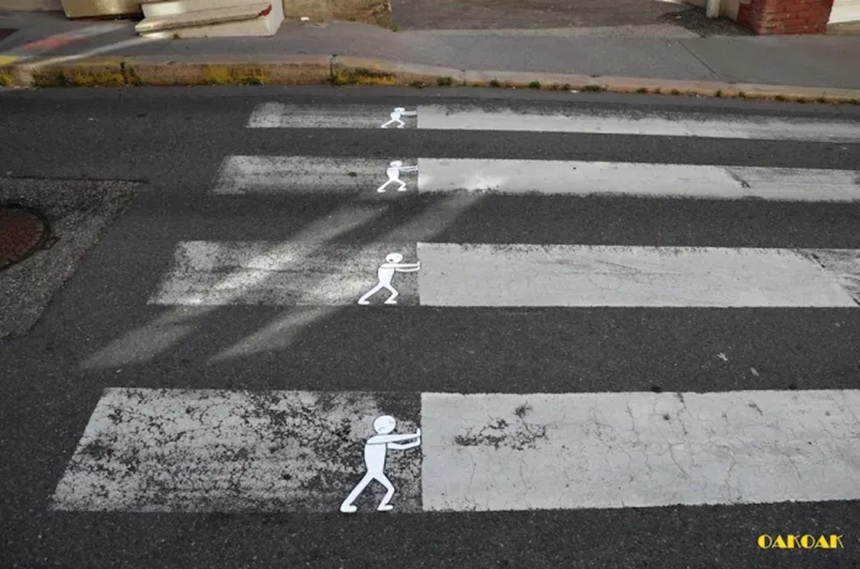 A street art intervention by Oakoak on a pedestrian crosswalk, depicting small white stick figures seemingly pushing the worn-out stripes of the crossing.