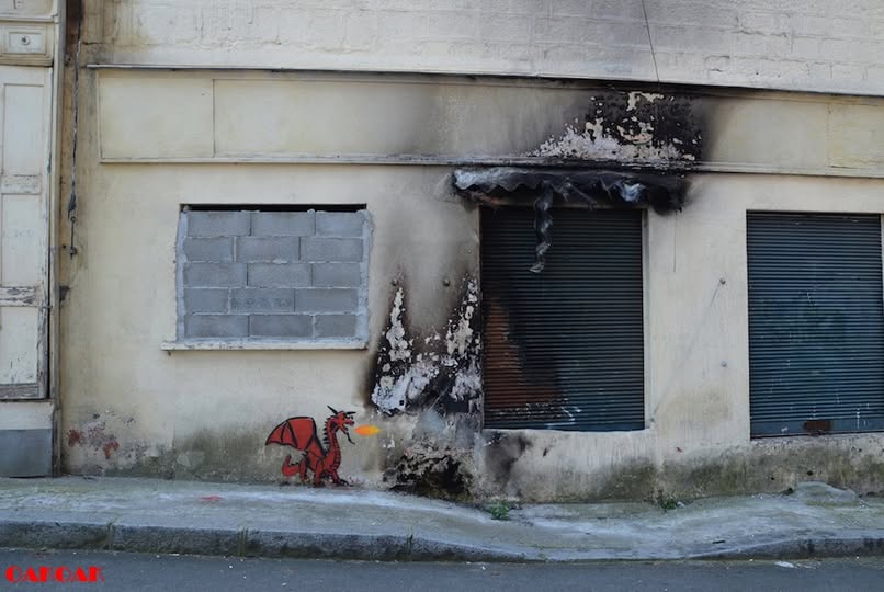 A clever street art piece by Oakoak incorporating fire damage on a building’s exterior. A small red dragon is painted near the burn marks, making it appear as though it has scorched the wall with its fiery breath.