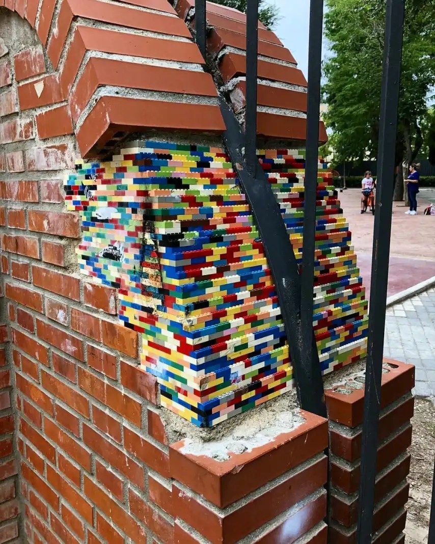 A crumbling red brick arch repaired with colorful LEGO bricks as part of Jan Vormann's Dispatchwork project. The vibrant LEGO patch fills the damaged area, contrasting sharply with the surrounding traditional bricks, while a black metal bar runs through the arch. In the background, a park with trees and people can be seen, adding an urban context to this playful intervention.