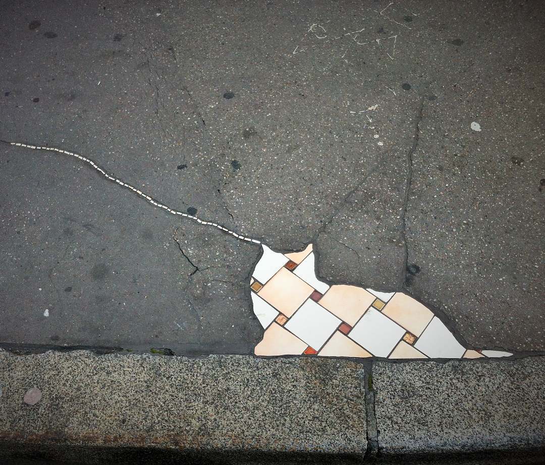 A playful mosaic by Ememem featuring a cat shape created from beige and orange tiles, integrated seamlessly into the pavement. The design uses a crack in the sidewalk to represent the cat’s tail, extending into the surrounding area. This whimsical artwork transforms urban damage into a charming and creative scene, inviting viewers to see beauty in imperfection.