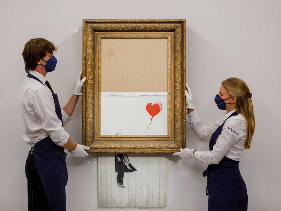 Two Sotheby’s staff members, wearing white gloves and blue masks, carefully hold Banksy's partially shredded artwork, formerly known as "Girl with Balloon," now titled "Love is in the Bin." The piece is encased in an ornate golden frame, with the bottom portion of the canvas shredded and hanging beneath. The upper part of the artwork features a red heart-shaped balloon floating against a blank background, emphasizing the contrast between creation and destruction. The scene is set in a gallery, highlighting the surreal and iconic nature of Banksy’s work.
