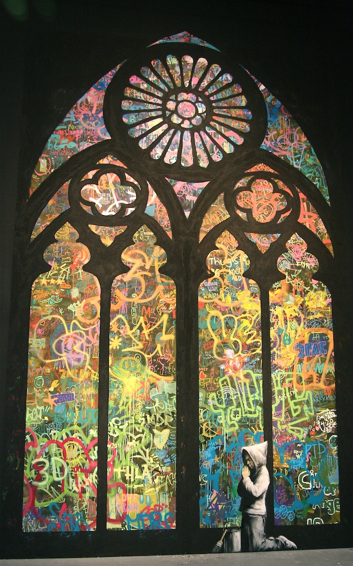 'Forgive Us Our Trespassing,' a large-scale mural created in 2011 by Banksy in collaboration with over 100 students from the City of Angels School. The artwork depicts a young figure in a hoodie, kneeling in prayer with hands clasped, positioned beneath a towering Gothic-style stained glass window filled with colorful graffiti. The vibrant, chaotic layering of tags, hearts, and peace signs contrasts with the solemn stance of the praying figure, blending religious iconography with urban street art to convey themes of forgiveness, rebellion, and the transformative power of creativity.