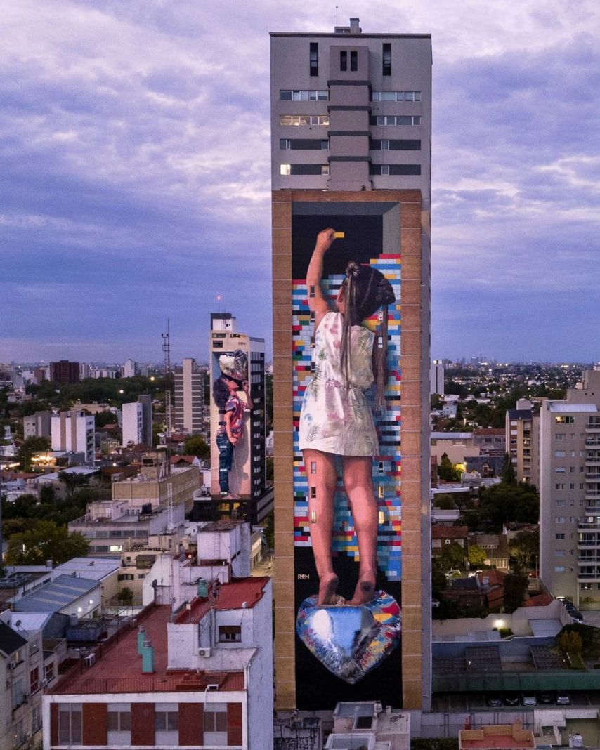 A monumental mural by Martín Ron in Banfield, Buenos Aires, Argentina. This artwork features a young girl in a floral dress standing barefoot on a reflective, heart-shaped sculpture. She reaches up with a yellow paint roller, seemingly painting a vibrant, pixelated background of multicolored blocks that extends across the tall building. The striking mural is part of an urban landscape, surrounded by city architecture under a pastel-hued evening sky. A smaller mural by Martín Ron can also be seen on a neighboring building in the background, adding depth to the scene.