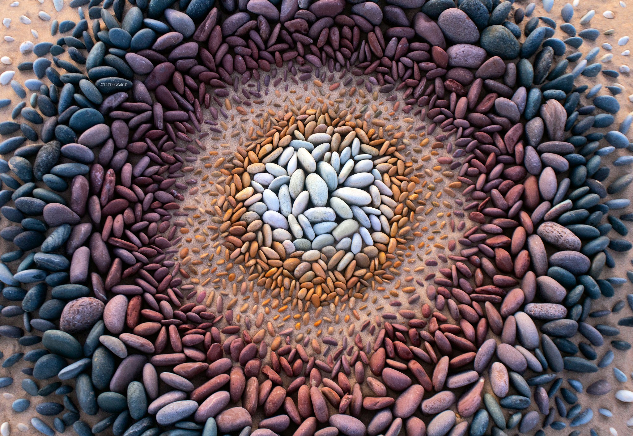 Dozens of tiny balanced stone stacks forming a symmetrical radial pattern on a pebble-covered beach, with an artist kneeling beside it.