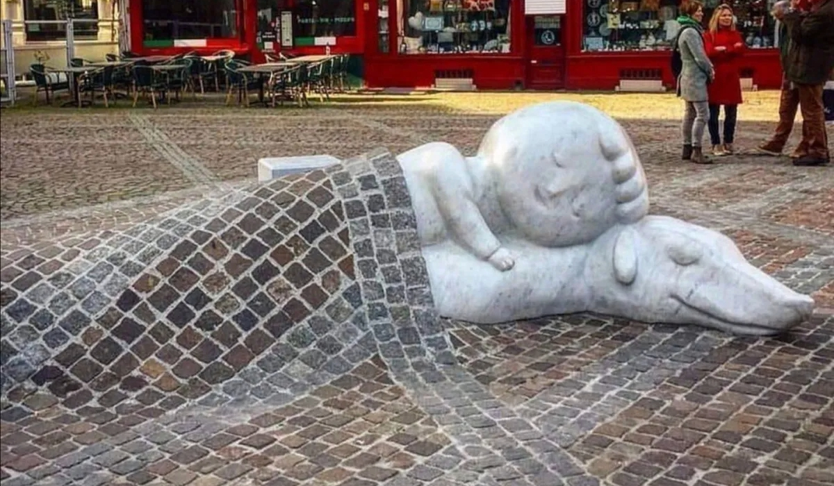 A touching sculpture of Nello and Patrasche, characters from the novel A Dog of Flanders, lying together under a cobblestone blanket. The artwork by Batist Vermeulen, also known as Tist, is located in Antwerp, Belgium, near the Cathedral of Our Lady. The sculpture symbolizes enduring friendship and resilience, set against a vibrant urban backdrop with historic architecture