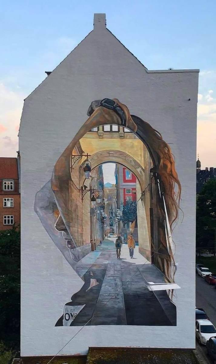 Large mural by Cristian Blanxer painted on the side of a building in Aarhus, Denmark, for the 17 Walls project. The artwork combines realism with surrealism, depicting the silhouette of a woman looking upwards. Inside her profile is a beautifully rendered street scene, as if viewing the scene through her mind’s eye. The detailed scene within the silhouette shows a European street with tall, historic buildings, arched architecture, and two pedestrians walking down a cobblestone path. The soft, warm lighting adds a sense of depth and tranquility to the piece, blending the woman's introspective expression with the urban landscape.