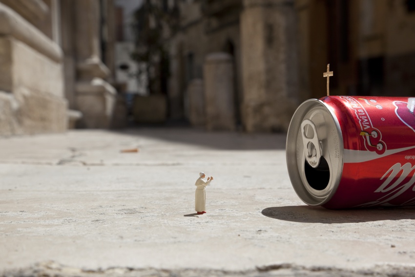 A piece from Slinkachu's 'Little People' series, featuring a miniature figurine of a priest or pope standing in front of a tipped-over soda can with a small cross planted on its top. The scene humorously resembles a religious moment or ceremony, with the soda can acting as an improvised altar. The background shows a blurred urban alley, contrasting with the playful foreground.