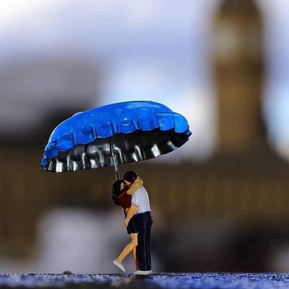 Close-up of two miniature figures embracing under a blue bottle cap used as an umbrella, with blurred urban architecture in the background.