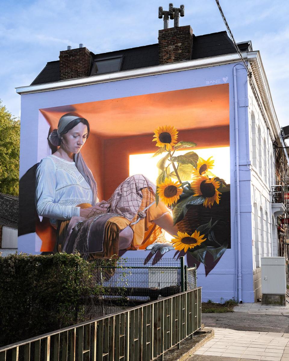 A captivating mural titled 'The Fisher Girl' by Fabian Bane Florin in Mons, Belgium. The artwork portrays a young woman sitting gracefully by a window, her hands delicately mending a fishing net. Sunflowers in vibrant yellow bloom nearby, basking in warm sunlight that floods the scene. The interplay of light and shadows creates a peaceful yet dynamic atmosphere. Painted on the side of a residential building, this mural celebrates themes of labor, nature, and serenity, blending realism with poetic storytelling.
