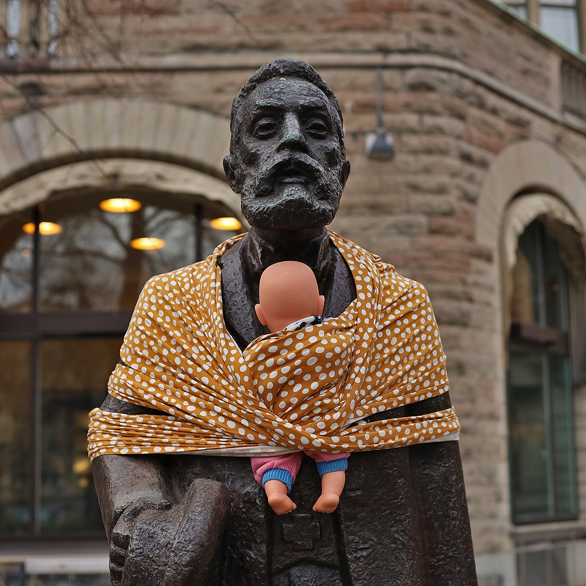A bronze statue of a historical figure is wrapped in a mustard-yellow baby sling with white polka dots, holding a doll dressed in colorful baby clothes. This creative modification is part of Arena Idé's International Men’s Day campaign to highlight unequal parenting roles in Sweden. The statue, set against a backdrop of stone architecture and softly glowing lights, symbolizes the campaign's call for increased paternal involvement in caregiving and shared parenting responsibilities. The use of the sling contrasts with the statue’s traditional stoic appearance, sparking thought and dialogue about modern fatherhood.