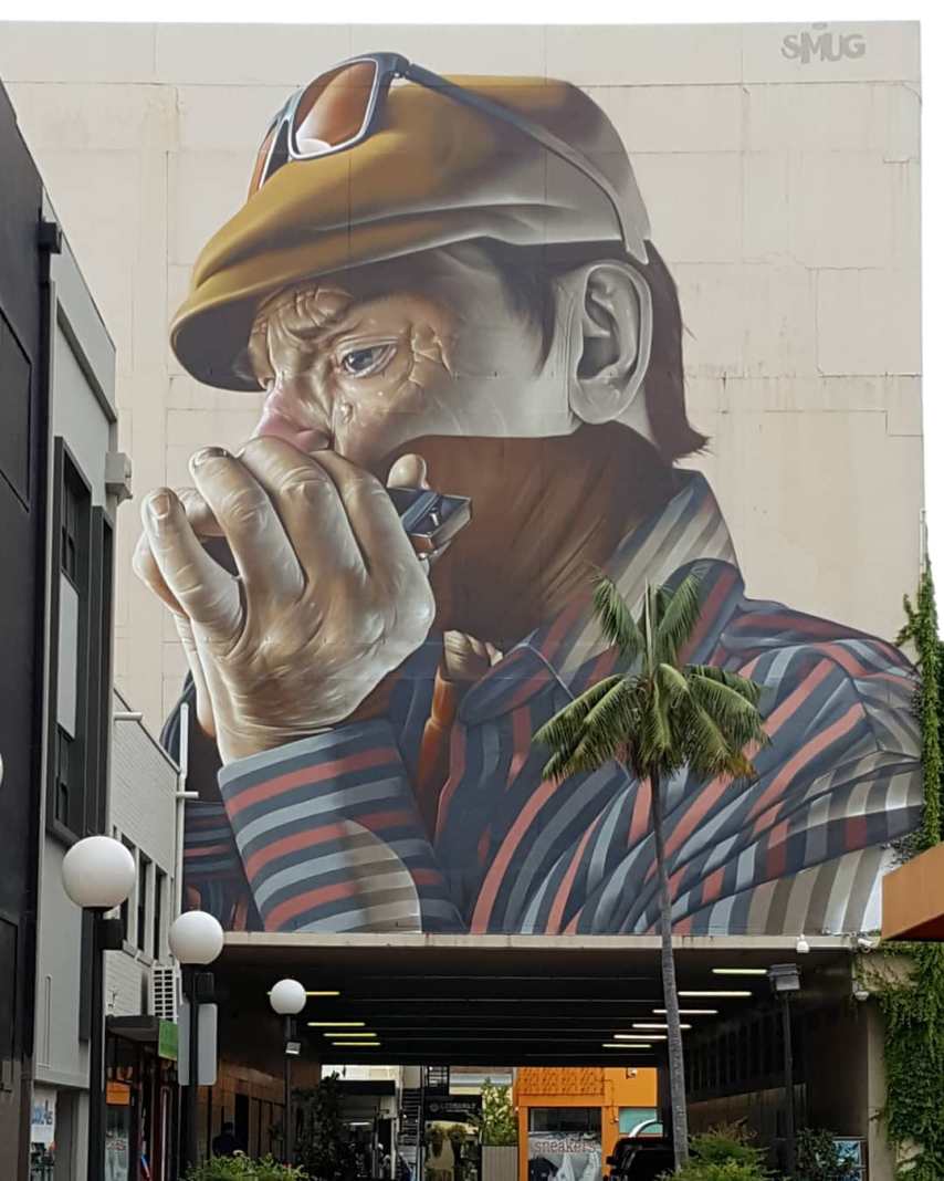 Mural by SMUG at the Wonderwalls Festival in Wollongong, Australia, depicting a close-up of a man playing a harmonica. The man wears a cap with sunglasses resting on top and a striped shirt, his face deeply focused on the music. The intricate details capture his facial wrinkles, expression, and the texture of his hands gripping the harmonica. This artwork, covering a large wall, showcases SMUG’s realistic style, with striking shading and color contrasts that bring the figure to life against the urban backdrop.