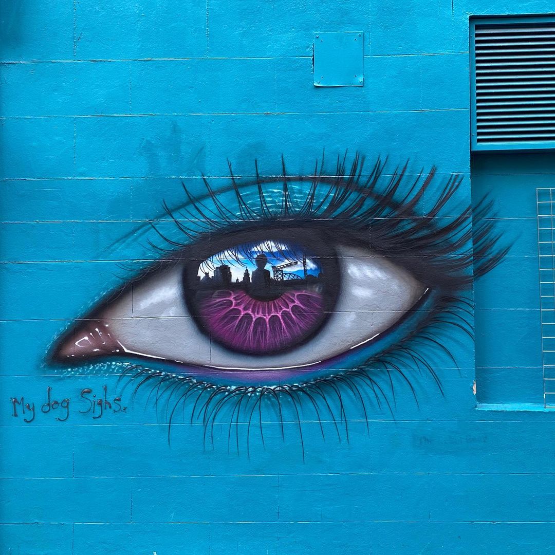 Eye mural by artist My Dog Sighs on a vibrant blue wall in Glasgow, Scotland. The mural features a striking eye with a deep purple iris, displaying intricate details and a reflective quality that mirrors the cityscape, including buildings and a partly cloudy sky. The eyelashes are long and detailed, adding to the realistic effect, while the pinkish-red tear duct contrasts with the bold blue background. The artist’s signature, 'My Dog Sighs,' is visible near the lower left corner, blending into the wall’s smooth surface. This mural, set against the bright blue wall, creates a powerful focal point with its surreal and expressive detail.