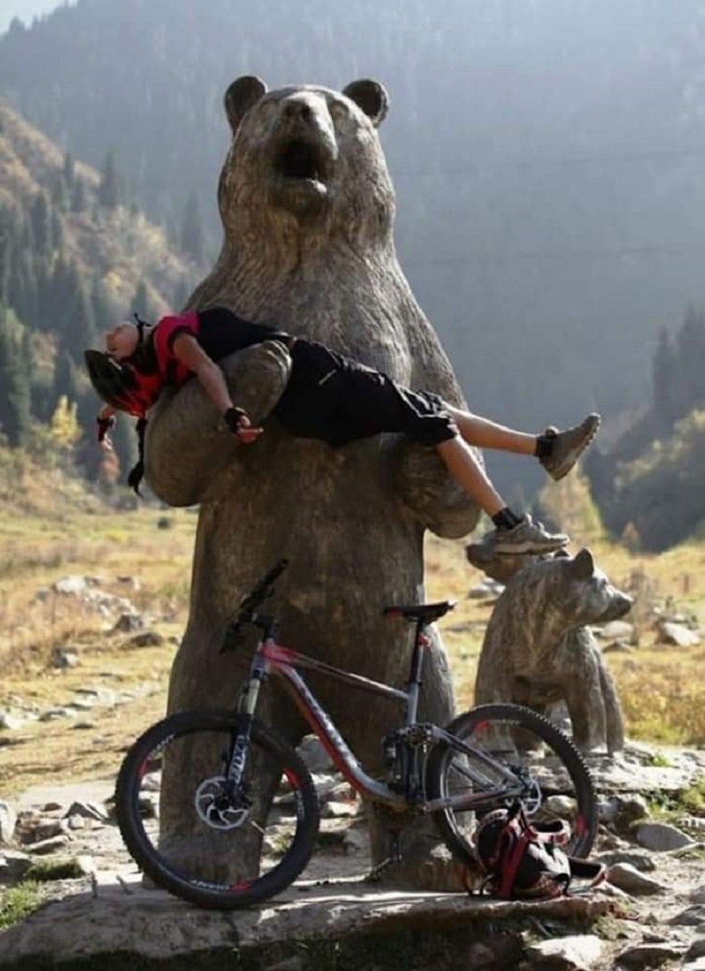 Mountain biker rests inside the arms of a giant wooden bear street art statue. A funny 3D illusion hug in the world of public graffiti and murals.