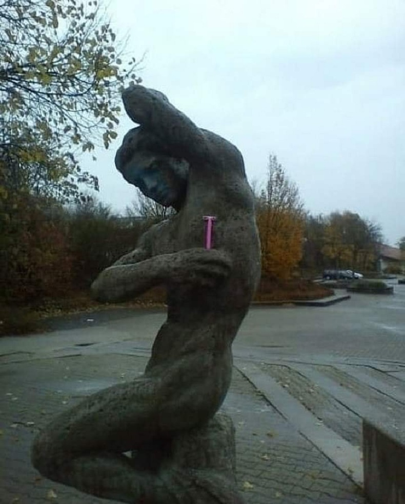 Humorous street art interaction featuring a pink razor placed under the arm of a classical Greek statue. A subtle 3D illusion and public graffiti intervention.